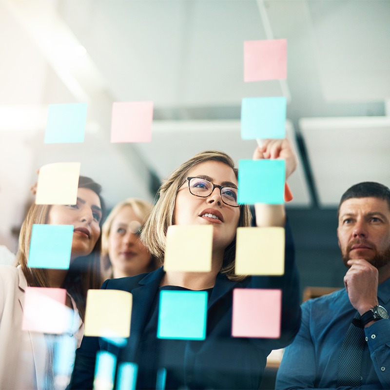 Mehrere junge Leute stehen hinter einer Glaswand und arbeiten an innovativen Ideen mit Post-its.